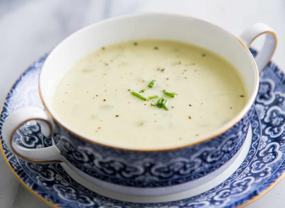 A bowl of creamy white soup garnished with chives and black pepper in a decorative blue and white cup and saucer. - Home Instead