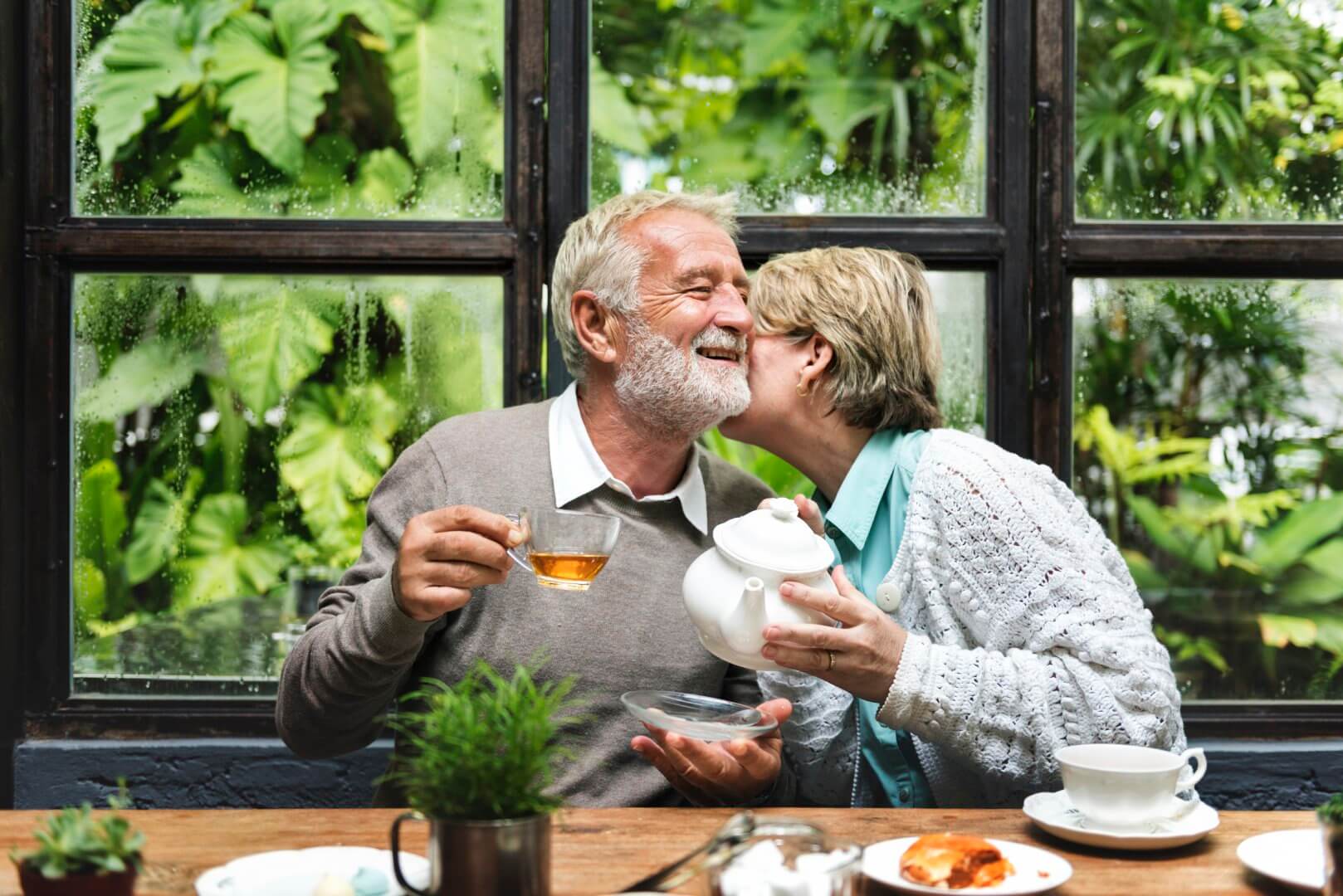 Elderly couple smiles and enjoys tea together in a cozy garden setting with lush greenery seen through the windows. - Home Instead