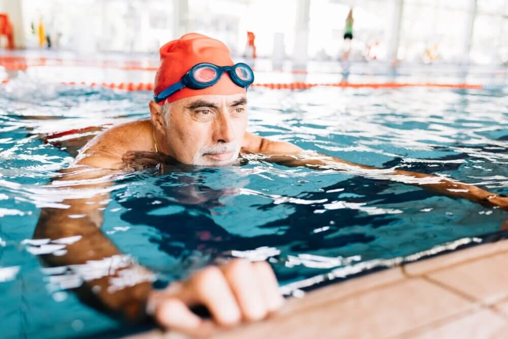 Elderly man wearing a red swim cap and blue goggles swimming in an indoor pool, holding the pool edge. - Home Instead