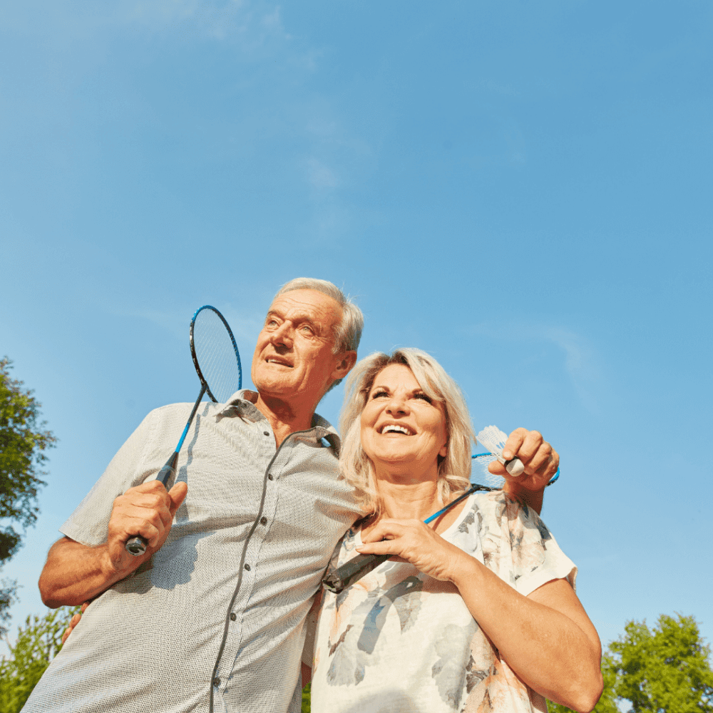 An elderly couple smiles and poses outdoors, holding badminton rackets and a shuttlecock under a clear blue sky. - Home Instead