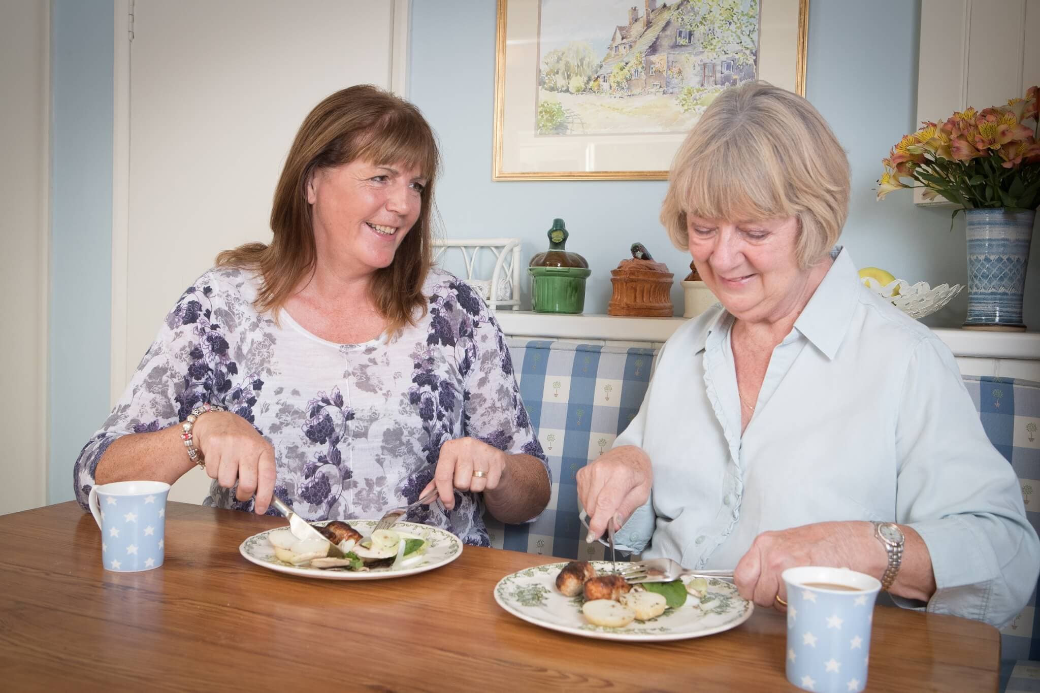Two women smiling and dining together at a table with plates of food and mugs in a cozy room. - Home Instead