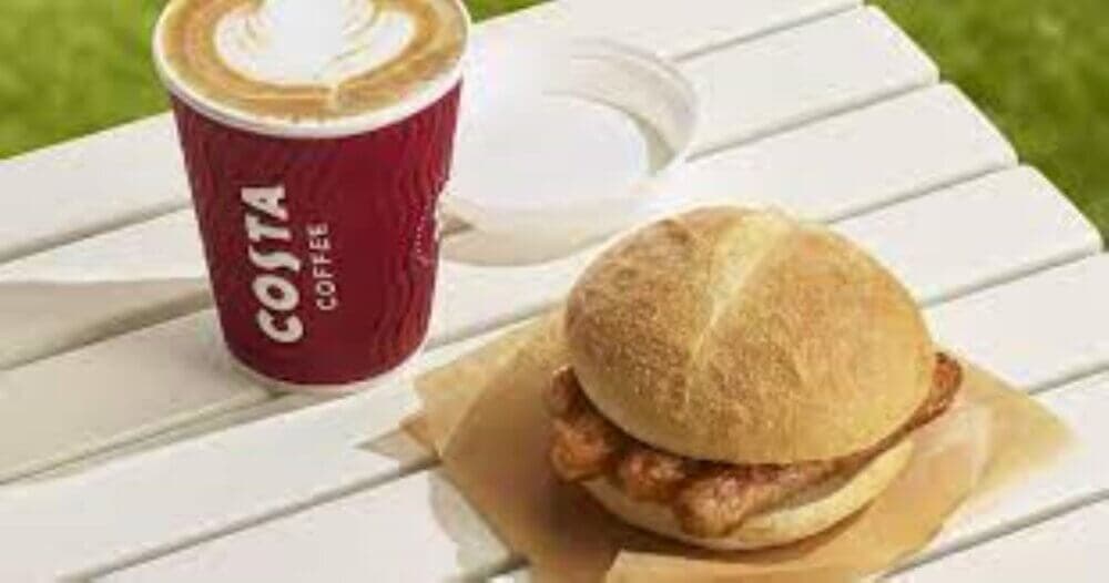 A Costa Coffee cup with a latte and a bacon sandwich on a white table outdoors. - Home Instead