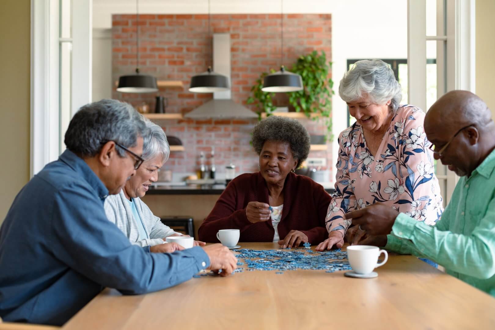 A group of older adults work on a jigsaw puzzle at a table, with coffee cups nearby in a cozy kitchen setting. - Home Instead