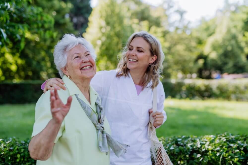 An elderly woman and a young woman, both smiling, walking and chatting in a park on a sunny day. - Home Instead