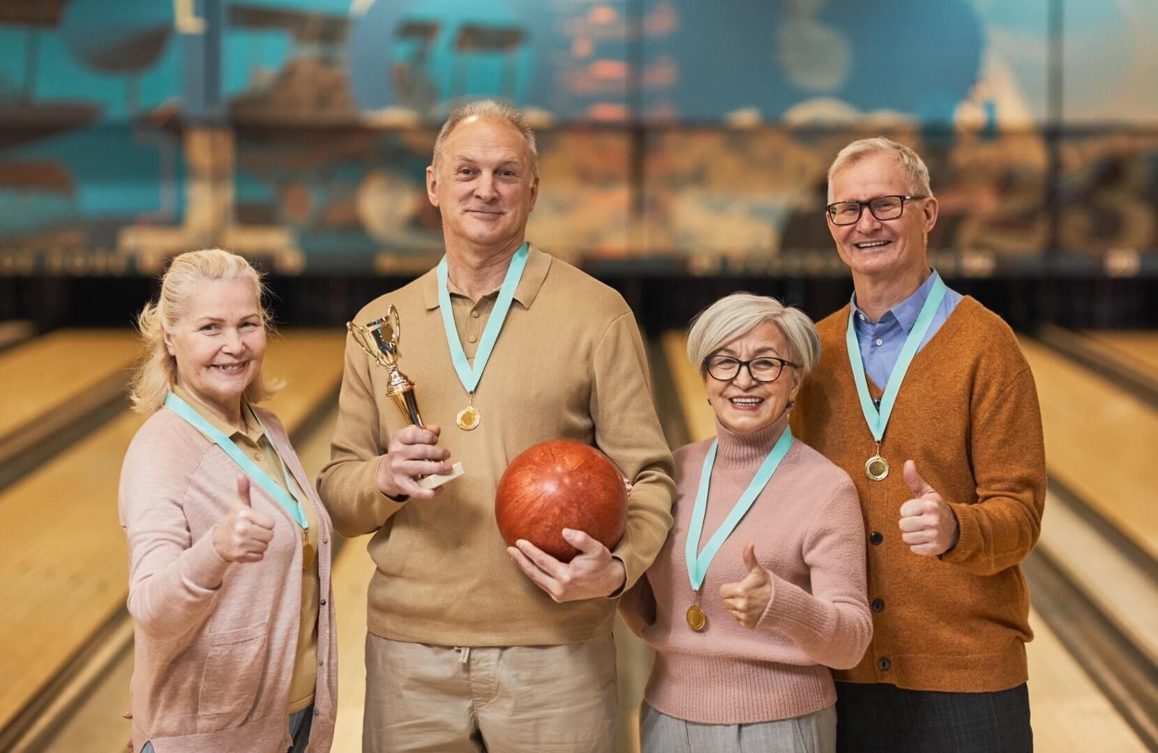 Four smiling seniors with medals pose in a bowling alley; one holds a trophy and a bowling ball. - Home Instead