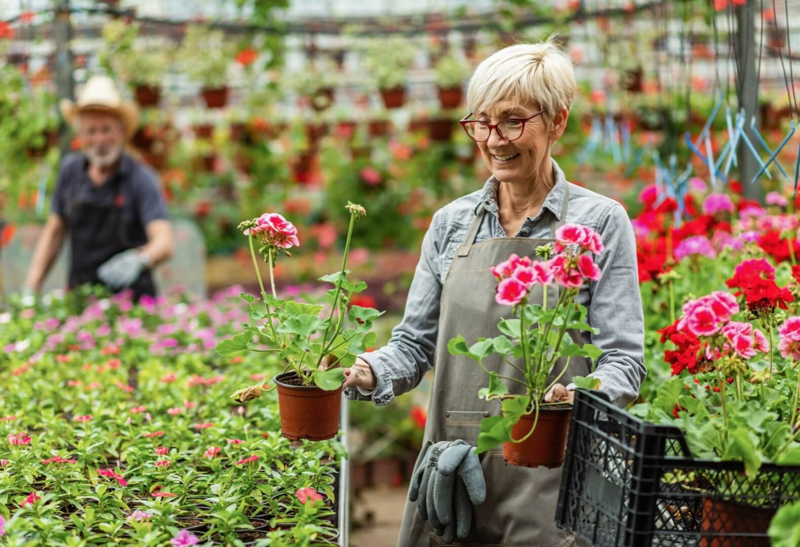 Woman with short blonde hair, glasses, and apron holds flower pots in greenhouse; man works in background among plants. - Home Instead