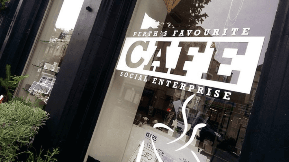 Storefront window displaying the text "Perth's Favourite Cafe Social Enterprise" with reflections of buildings. - Home Instead