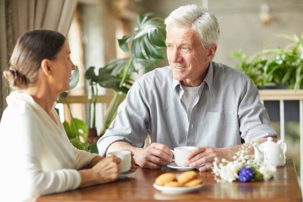 Two elderly people enjoying coffee and conversation at a table with cookies and flowers in a cozy setting. - Home Instead