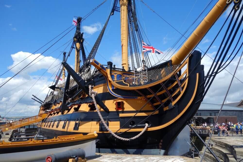 A historic sailing ship with intricate rigging and a prominent British flag docked at a sunny harbor. - Home Instead