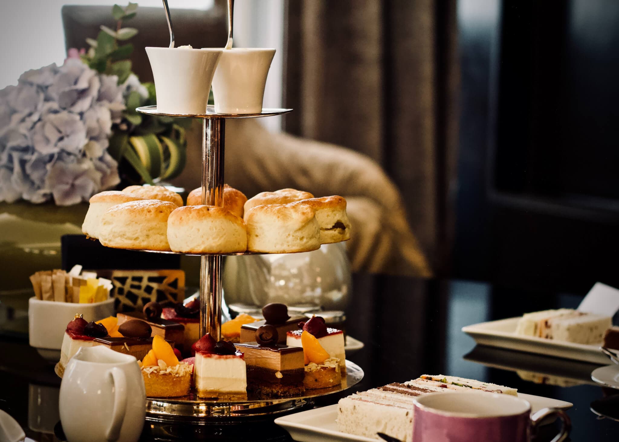 Two-tiered tray with scones, pastries, and desserts, surrounded by tea cups and a teapot on a table. - Home Instead