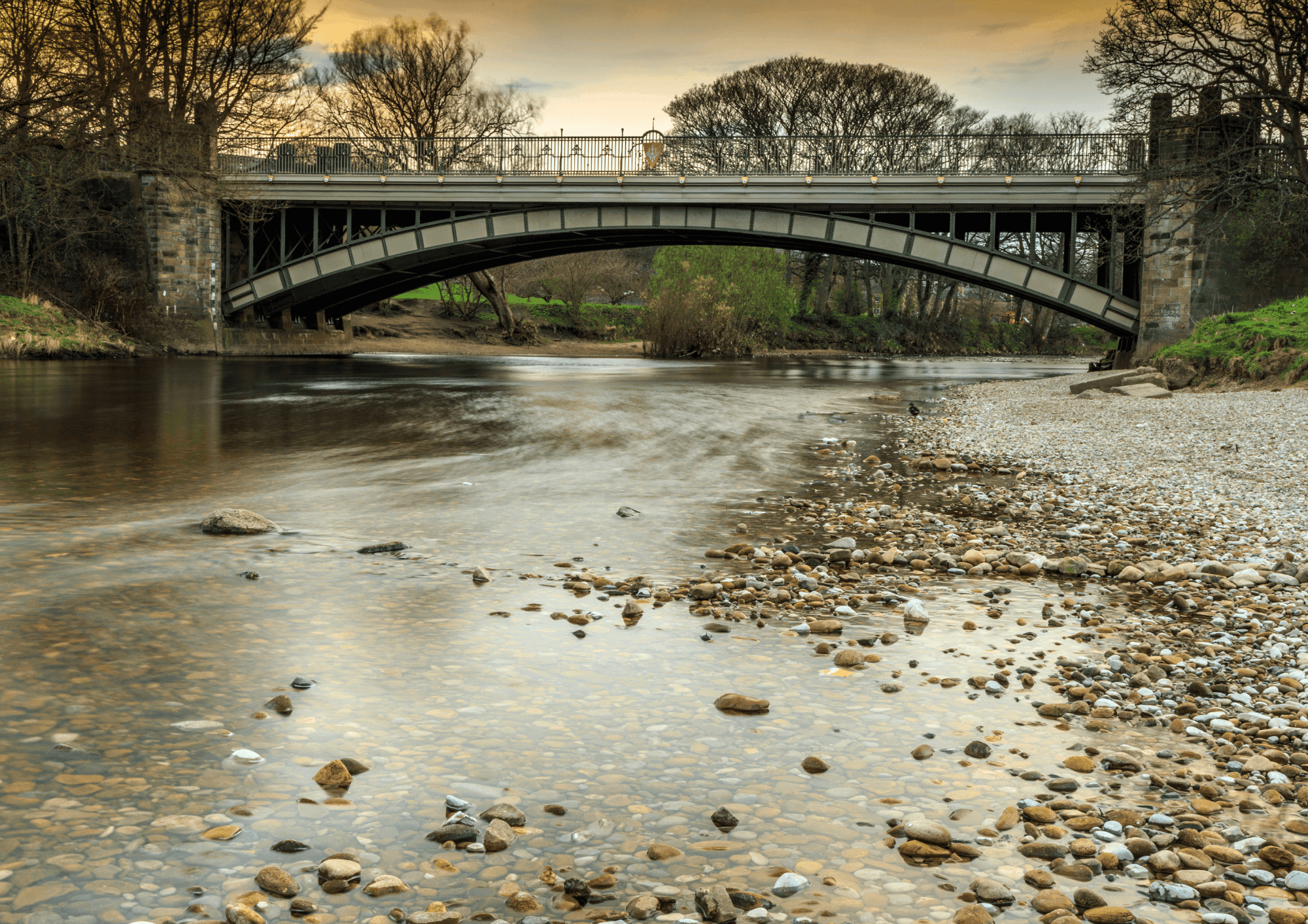 A serene river with a pebble-strewn shore flows under a stone arch bridge at dusk, with bare trees in the background. - Home Instead
