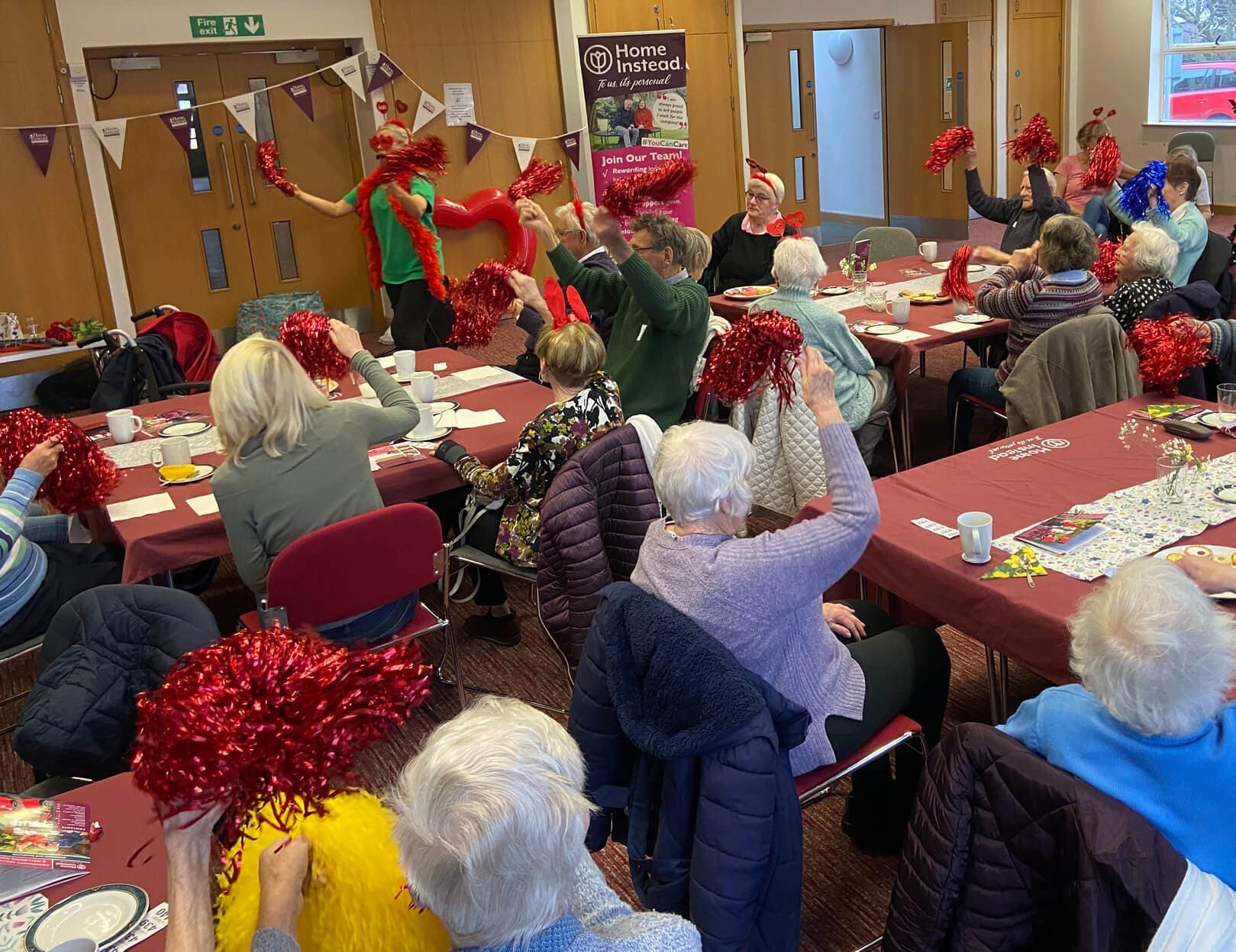 A group of seniors seated around tables raising red pom-poms during a festive indoor event. - Home Instead