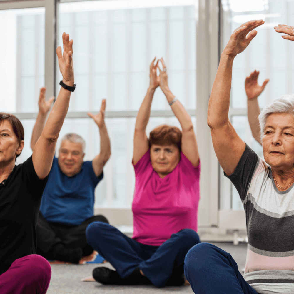 A group of elderly individuals practicing seated yoga with their arms raised in a bright room. - Home Instead