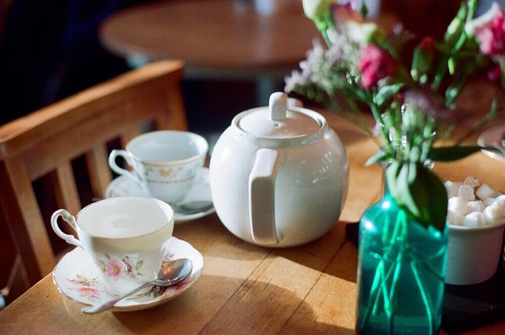 A wooden table with a teapot, two teacups, a vase of flowers, and a bowl of sugar cubes. - Home Instead
