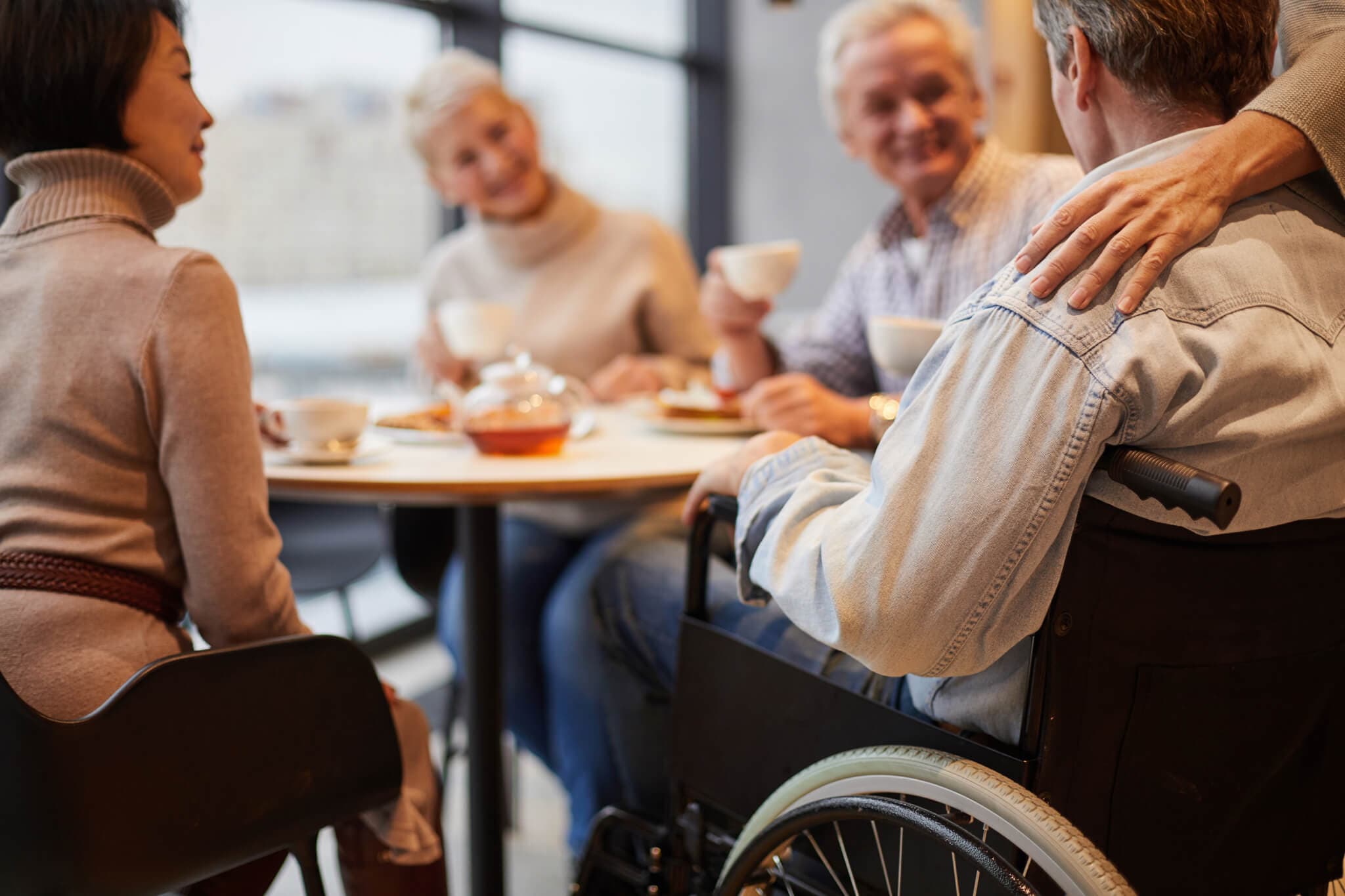 Group of people sitting at a café table, one in a wheelchair, chatting and enjoying tea. A hand rests on the wheelchair. - Home Instead