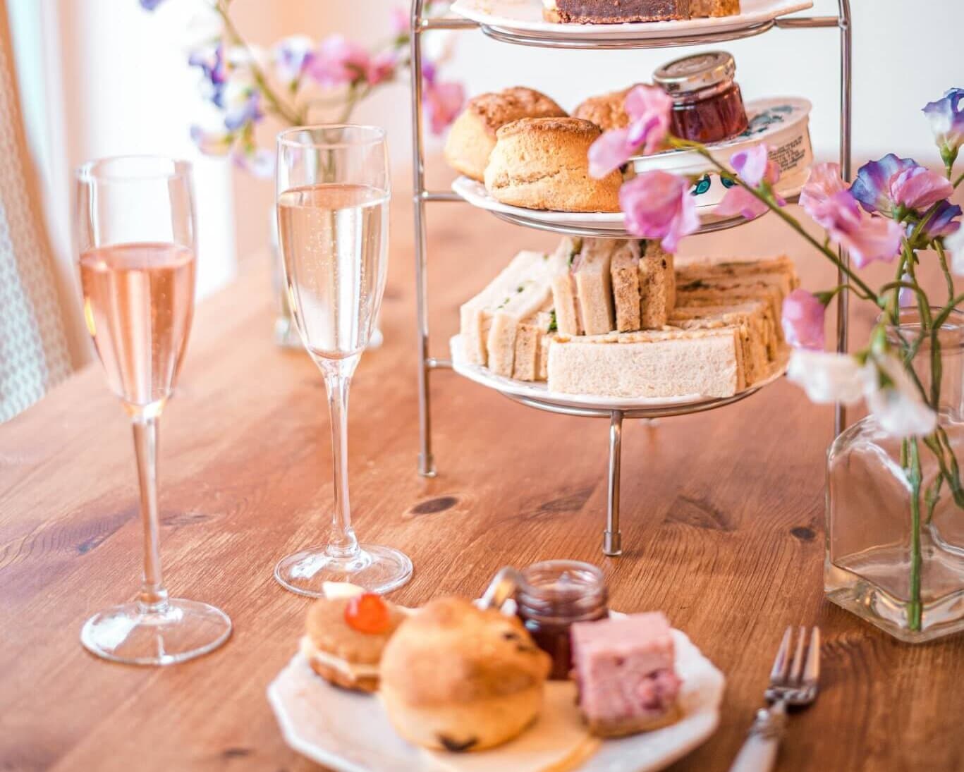 A tiered stand with pastries and sandwiches, flanked by champagne glasses and flowers on a wooden table. - Home Instead