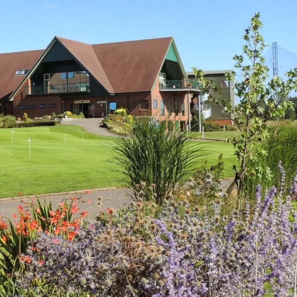 A clubhouse with a sloped roof and balcony overlooks a golf course, surrounded by colorful flowers and greenery. - Home Instead