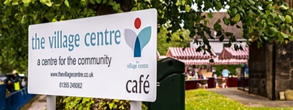 Sign for "The Village Centre" with a café, surrounded by trees, with a market stall visible in the background. - Home Instead