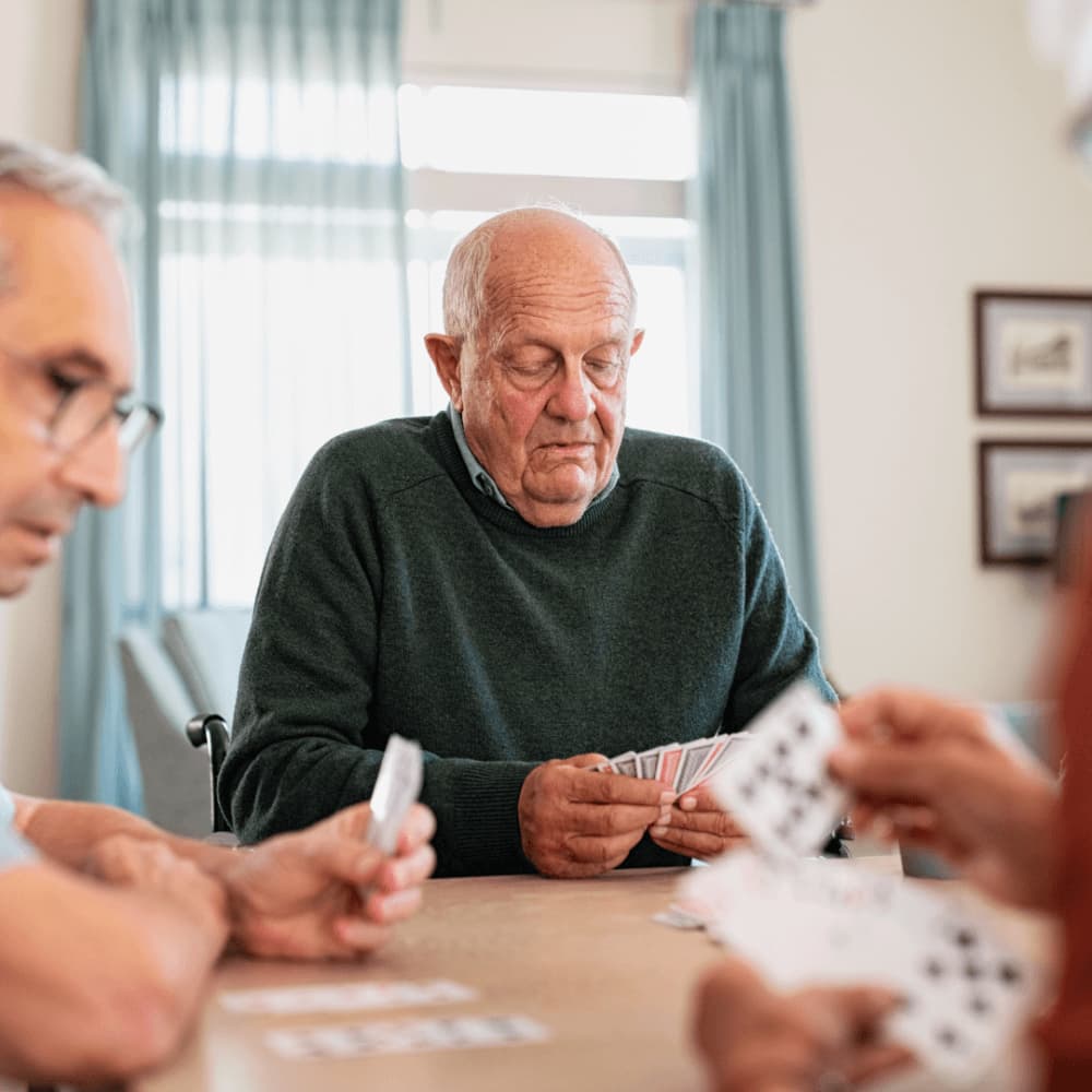 Three elderly individuals play cards together at a table in a bright room with curtains and framed pictures in the background. - Home Instead