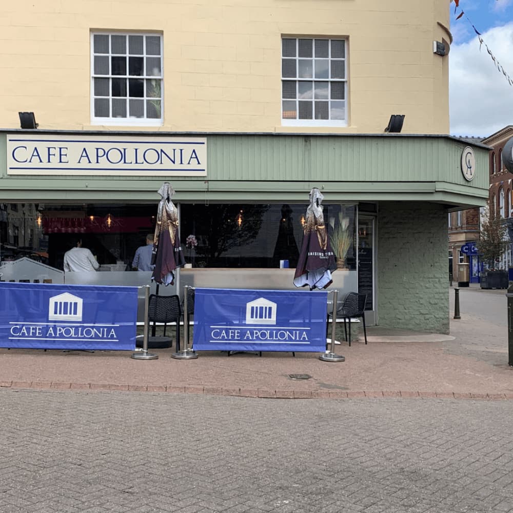 Outdoor seating and front entrance of Cafe Apollonia in a light yellow building on a brick-paved street. - Home Instead