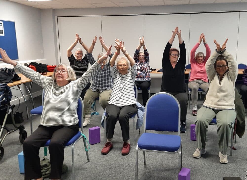 A group of older adults sitting on chairs, raising their arms overhead in a group exercise or yoga class. - Home Instead