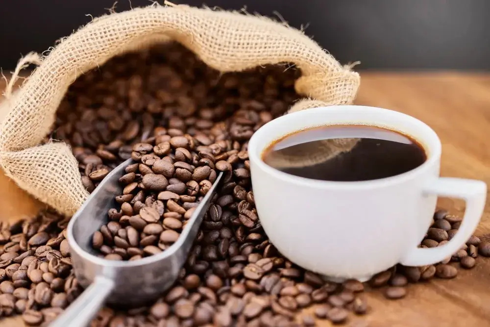 A cup of black coffee next to a burlap sack with spilled coffee beans and a metal scoop on a wooden surface. - Home Instead