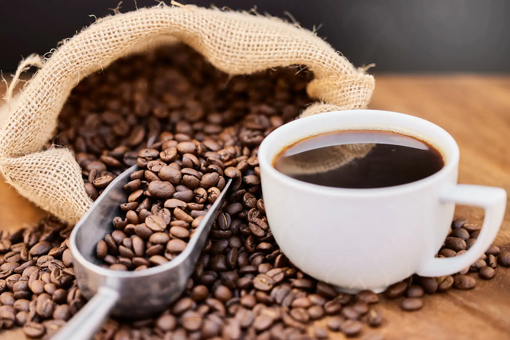 A cup of black coffee next to a burlap sack with spilled coffee beans and a metal scoop on a wooden surface. - Home Instead