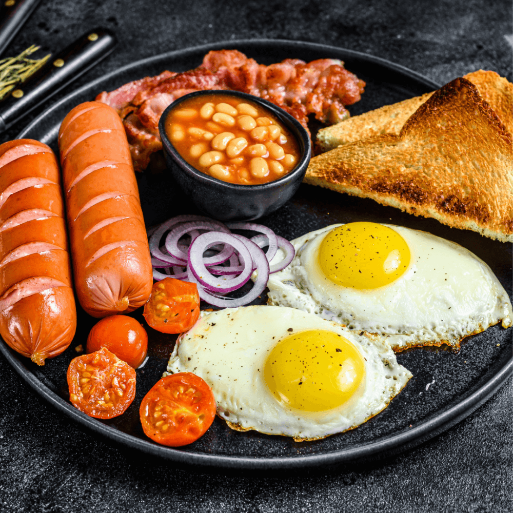 A full English breakfast with fried eggs, sausages, tomatoes, baked beans, toast, bacon, and sliced onions on a black plate. - Home Instead