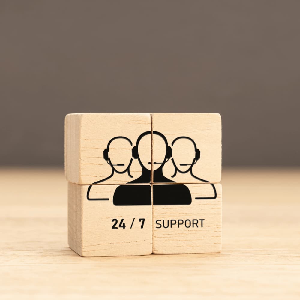 Wooden blocks with an icon of three customer service representatives with headsets and the text "24 / 7 SUPPORT. - Home Instead