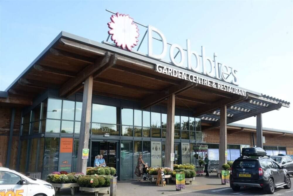 Exterior of Dobbies Garden Center & Restaurant with plants, people, and parked cars in front. - Home Instead