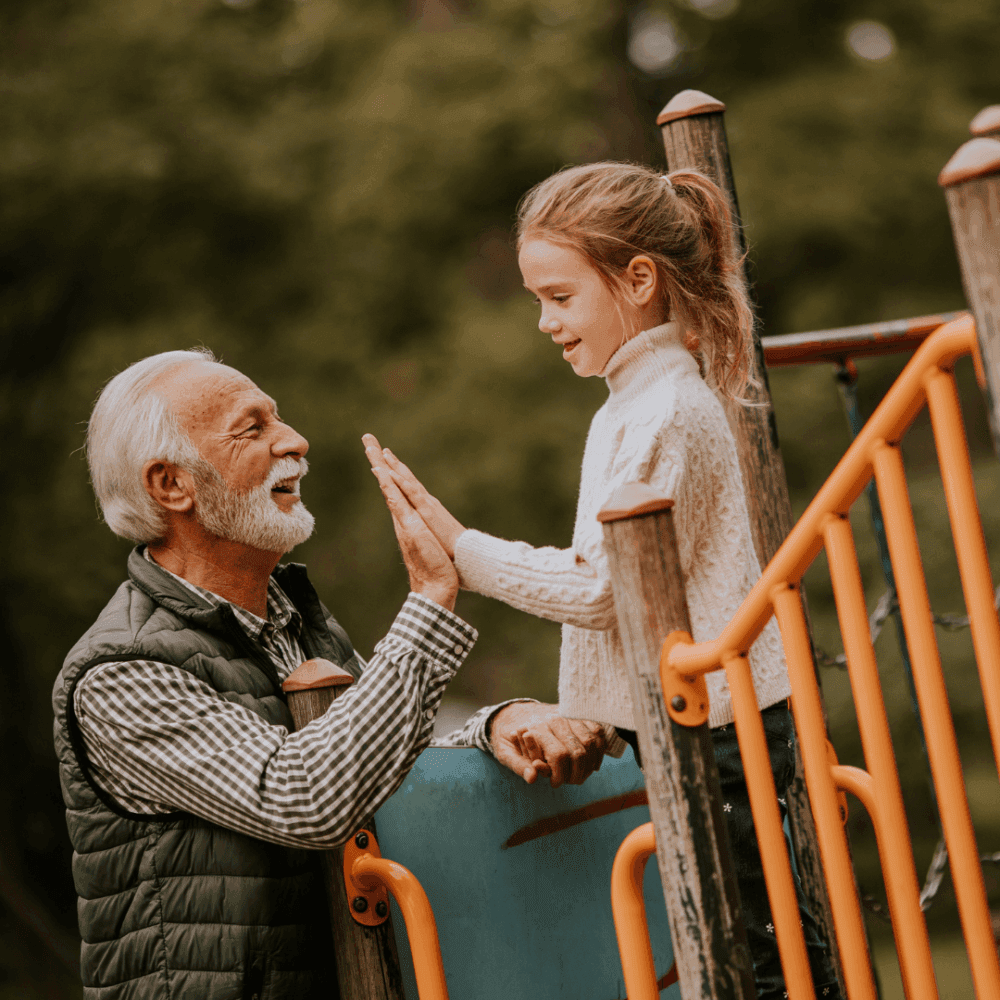 A bearded man and a young girl share a high-five on a playground with green trees in the background. - Home Instead
