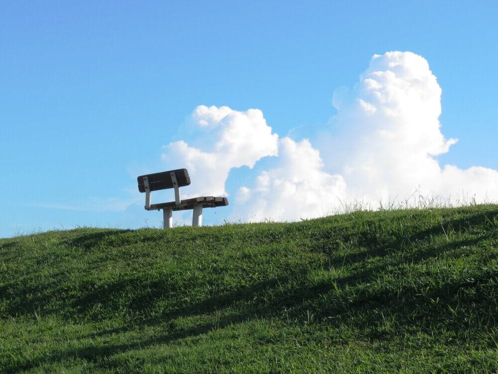 A solitary park bench sits atop a grassy hill under a bright blue sky with fluffy white clouds. - Home Instead