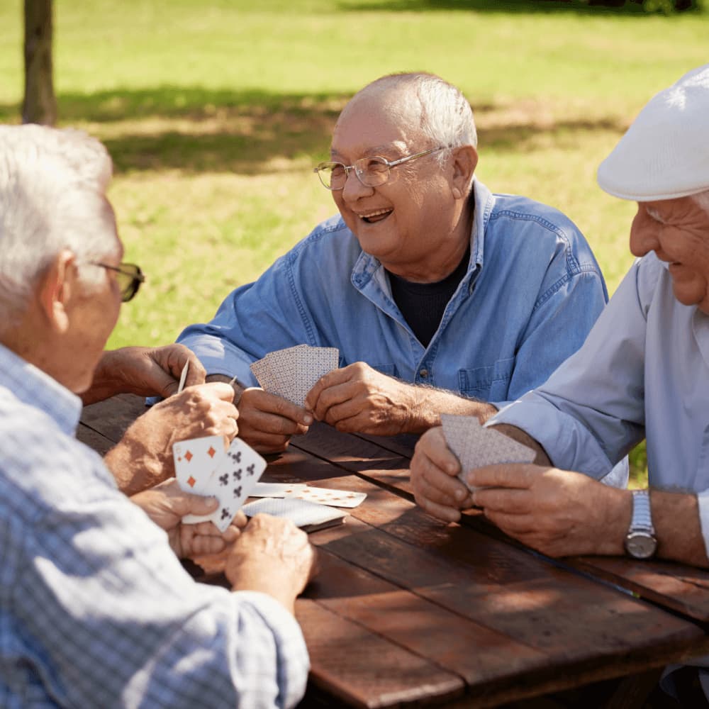 Four older men enjoying a card game outdoors, smiling and laughing around a picnic table in a sunny park. - Home Instead