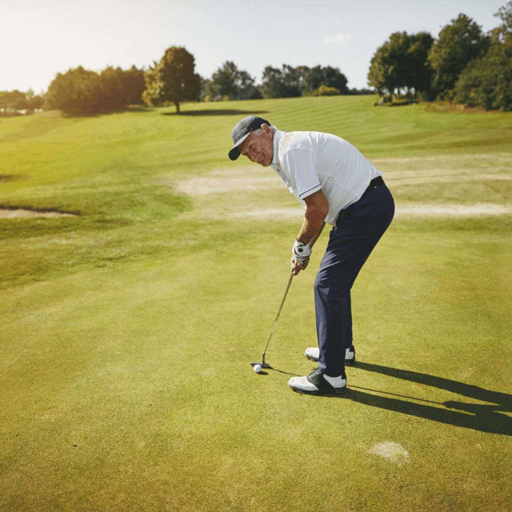 A man in a white shirt and blue pants, standing on a golf course, about to putt a golf ball with his club. - Home Instead