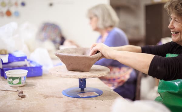 Two people shape clay on pottery wheels, focusing on creating ceramic pieces in a studio setting. - Home Instead