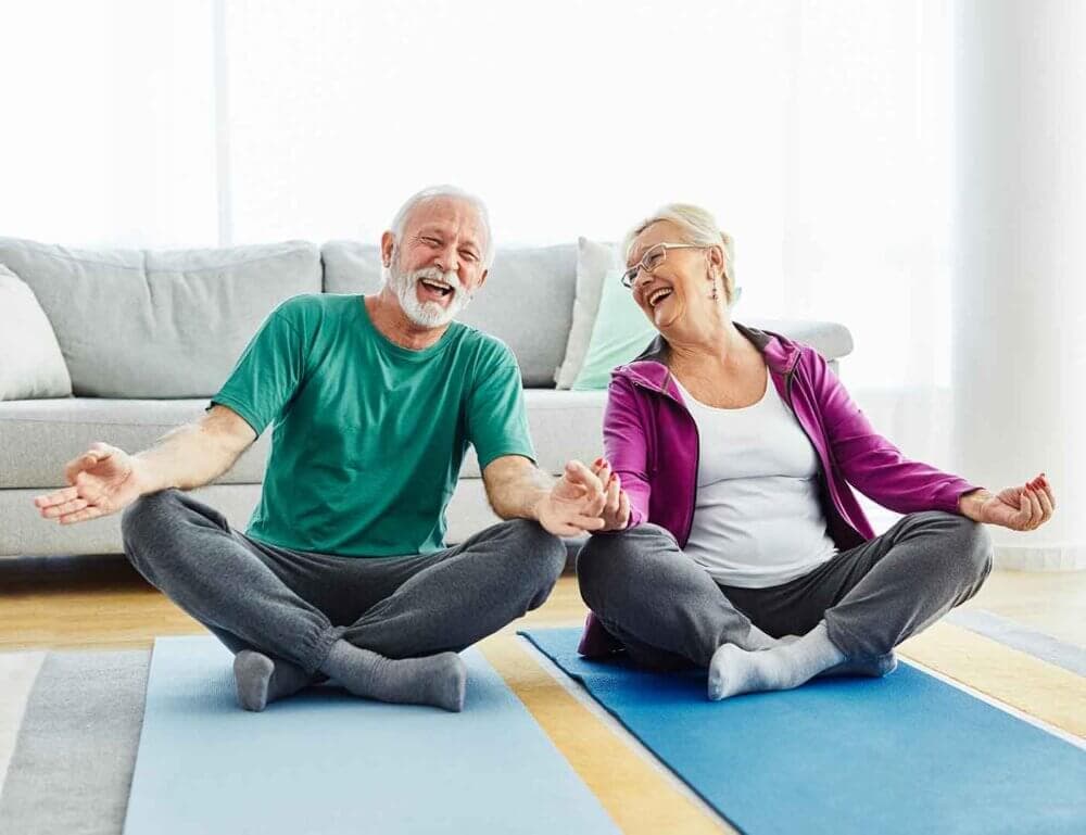 Elderly couple laughing and sitting in lotus position on yoga mats in a bright room. - Home Instead