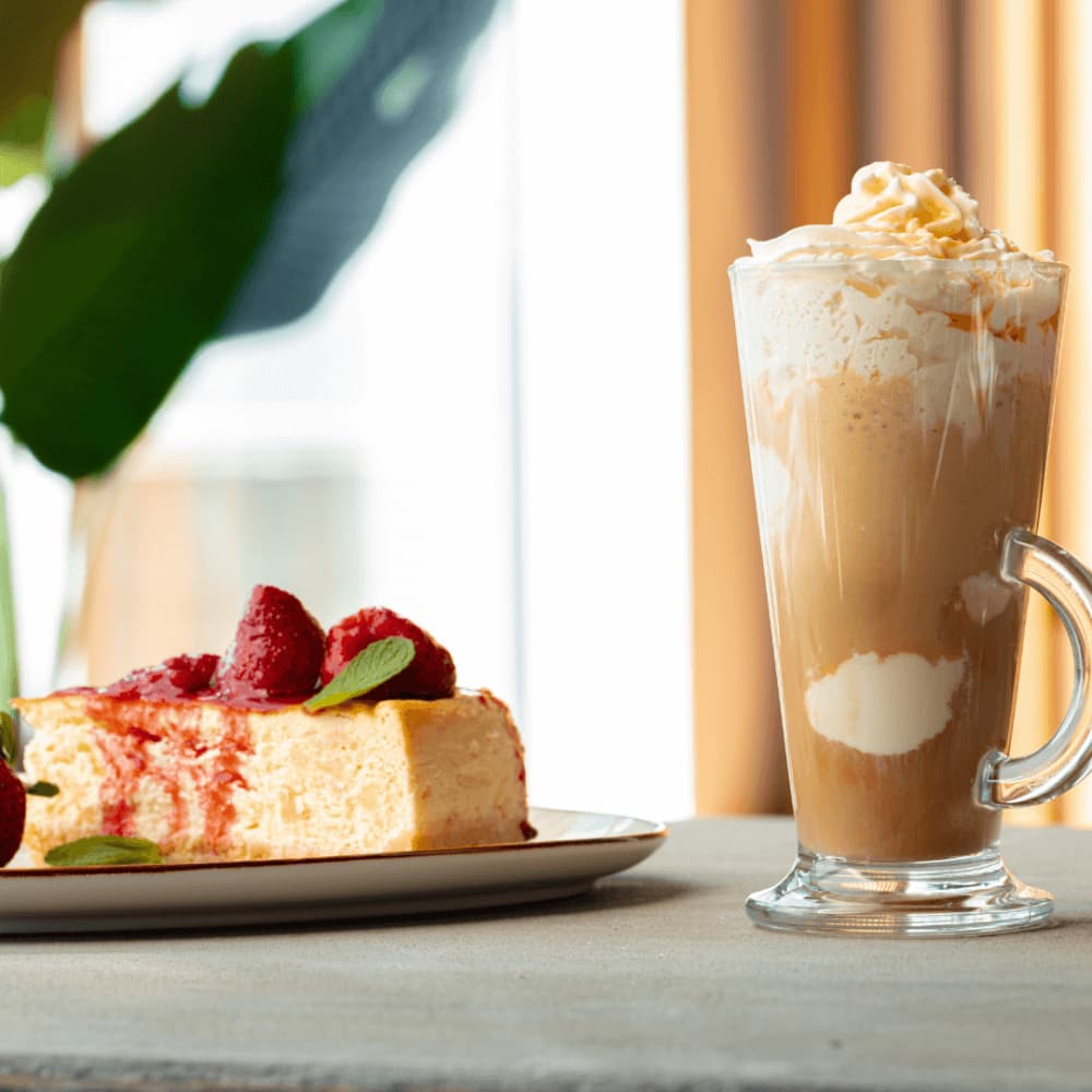 Slice of cheesecake with raspberries on a plate and a tall glass of iced coffee topped with whipped cream on a table. - Home Instead