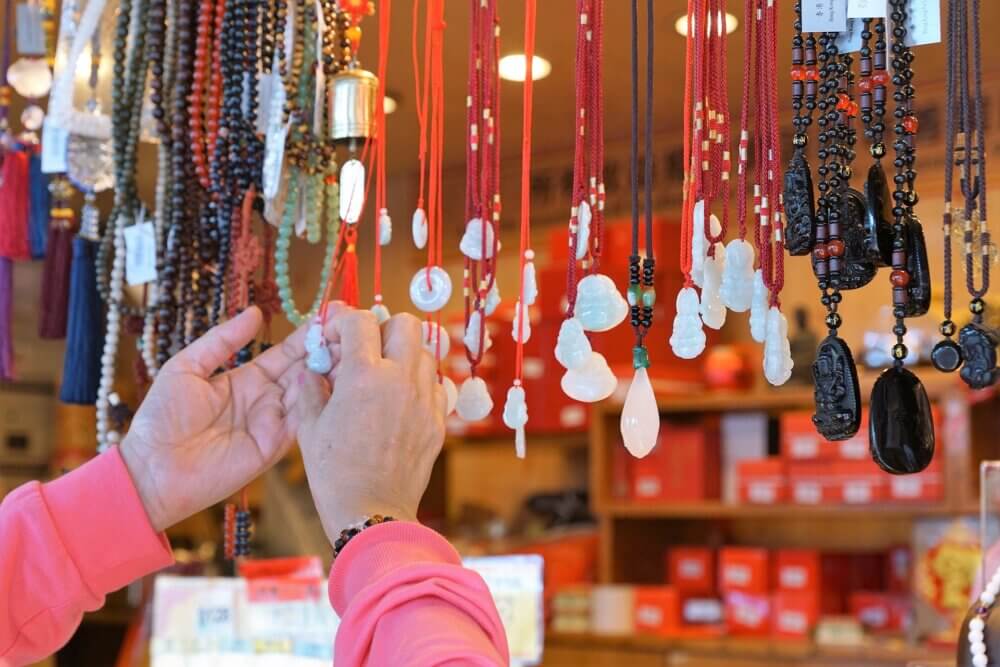 A person in a pink sleeve holds decorative necklaces with white and black pendants at a shop with red shelves. - Home Instead