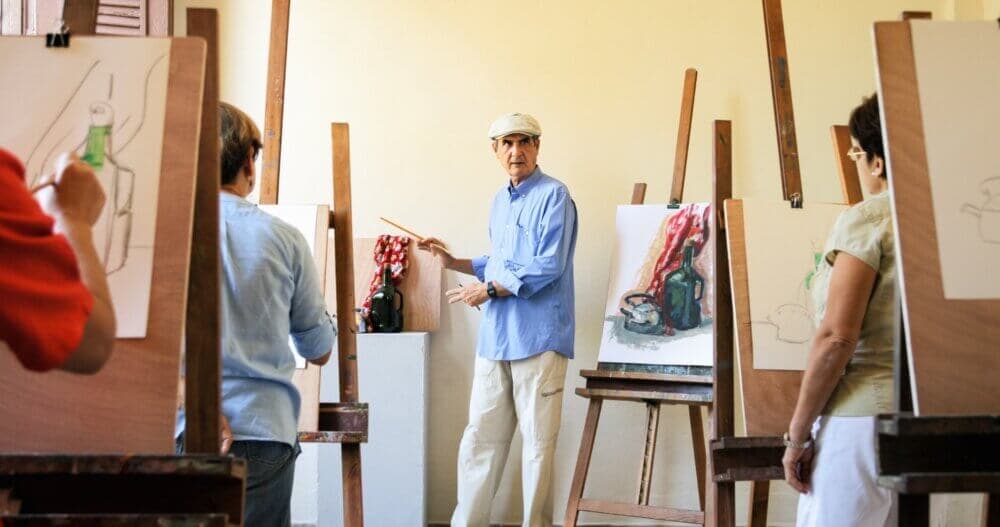 An art instructor demonstrates in front of easels while students observe and sketch in a classroom setting. - Home Instead