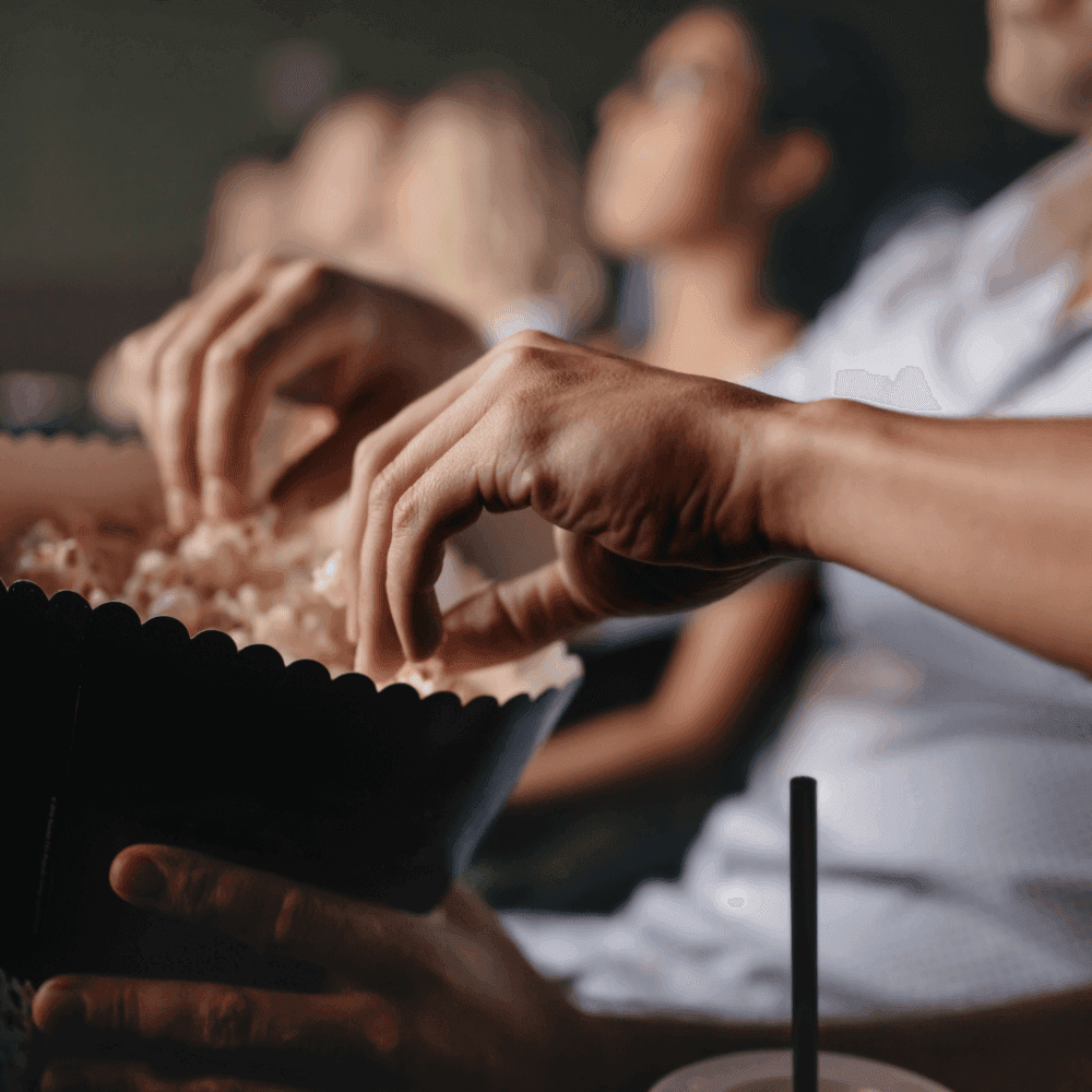 Close-up of hands reaching into a popcorn bucket in a movie theater with blurred audience in the background. - Home Instead