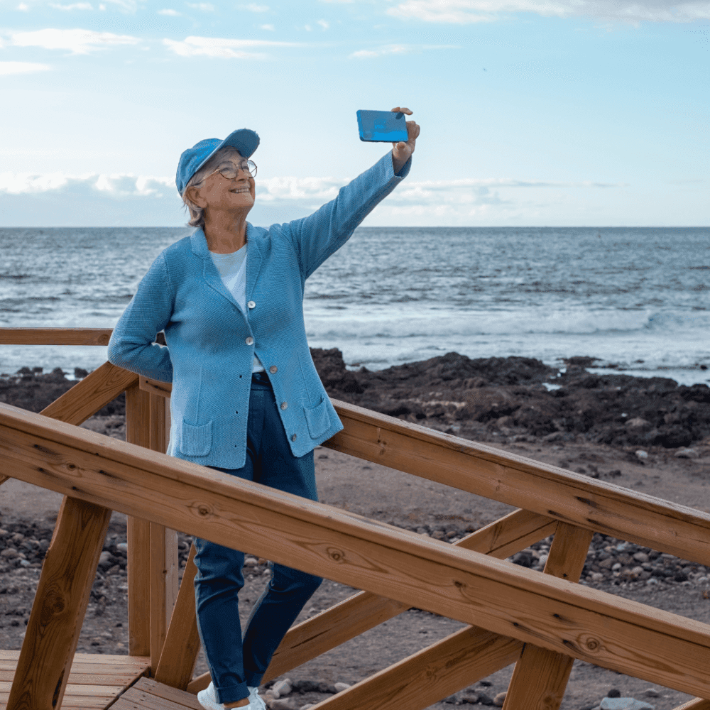 Elderly woman in blue outfit taking a selfie by the ocean, standing on a wooden boardwalk with a bright sky in the background. - Home Instead