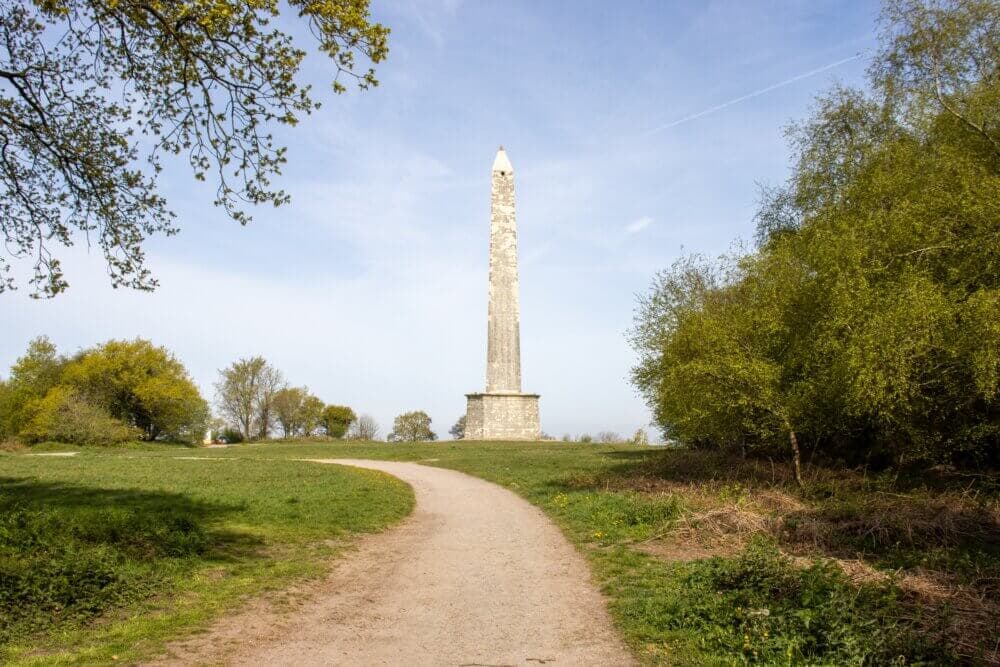 A tall stone obelisk stands on a grassy hill surrounded by trees, with a dirt path leading up to it under a blue sky. - Home Instead