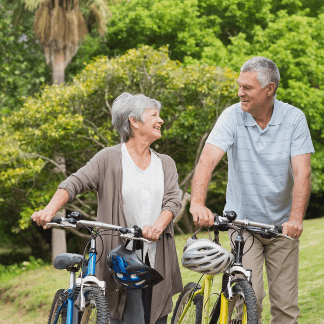 An elderly couple smiling at each other while holding bicycles in a lush green park. - Home Instead