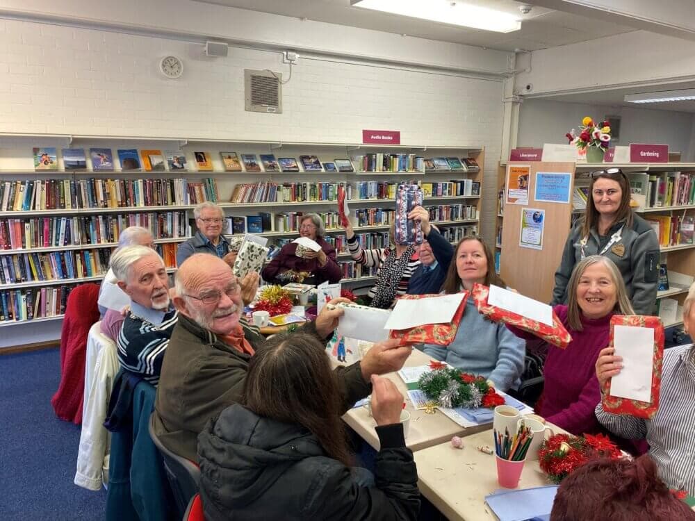 A group of people in a library celebrating with gifts and decorations around a table, smiling and holding presents. - Home Instead