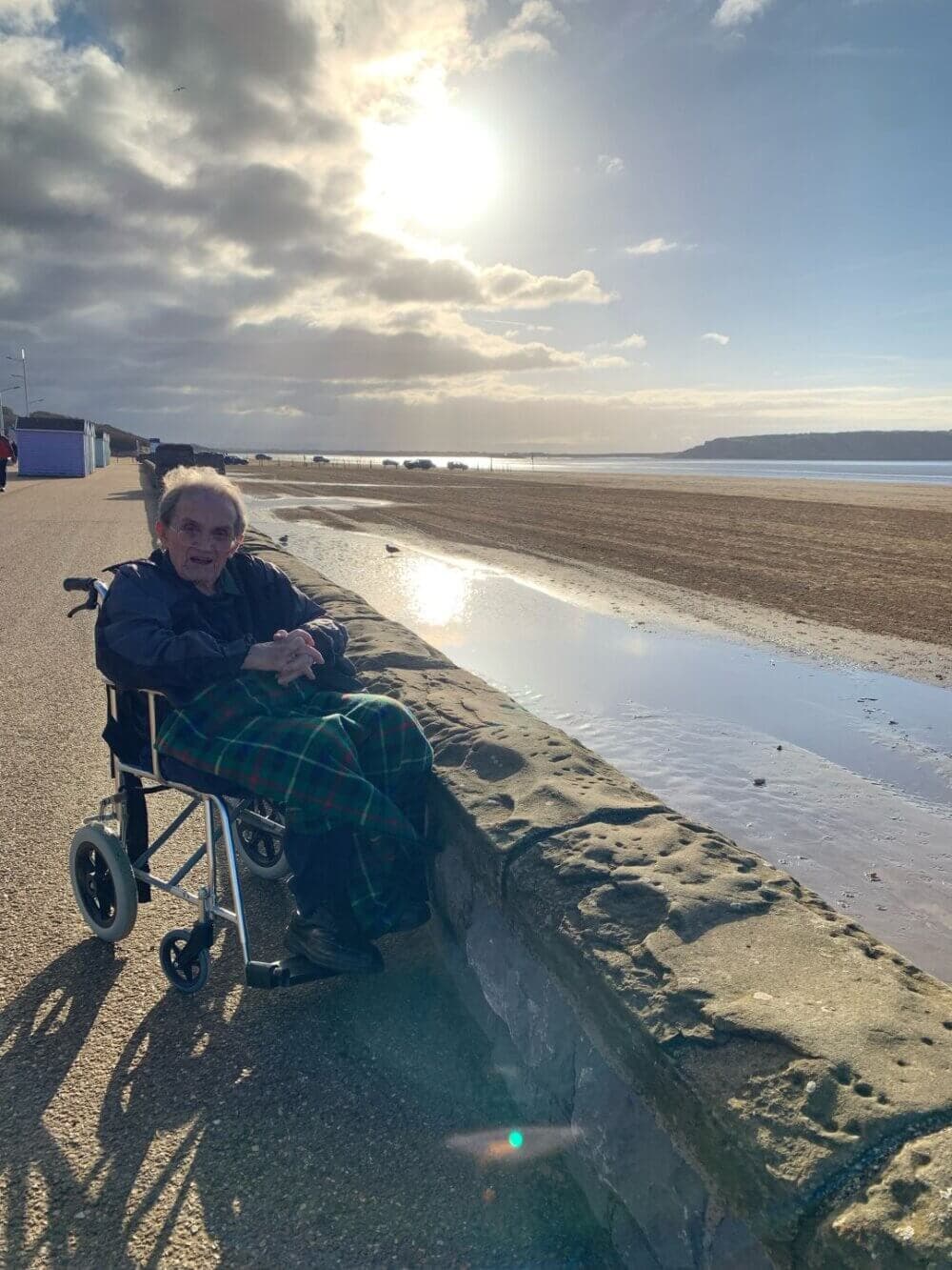 An elderly person in a wheelchair enjoys a sunny day by the seaside with a sandy beach and cloudy sky in the background. - Home Instead