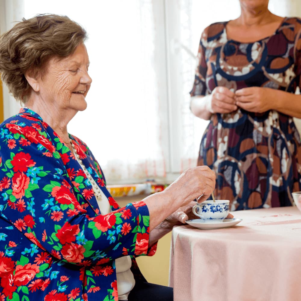 Elderly woman in a floral jacket stirs a cup of tea while seated at a table; another person stands nearby. - Home Instead