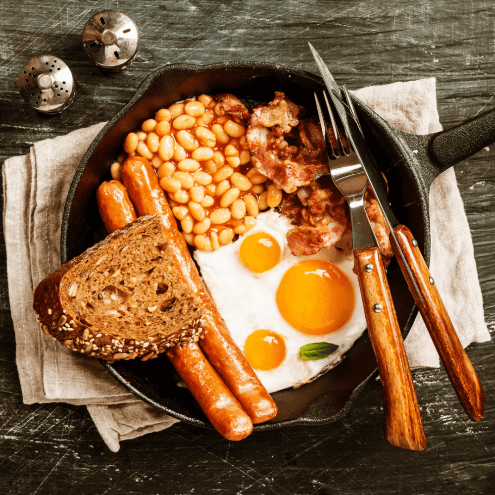 A skillet breakfast with fried eggs, sausages, baked beans, bacon, and toast, with cutlery placed on a linen napkin. - Home Instead