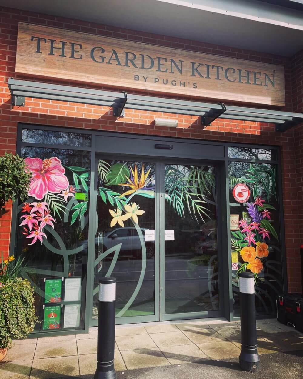 Entrance of The Garden Kitchen with vibrant flower mural on glass doors and sign above, framed by greenery. - Home Instead