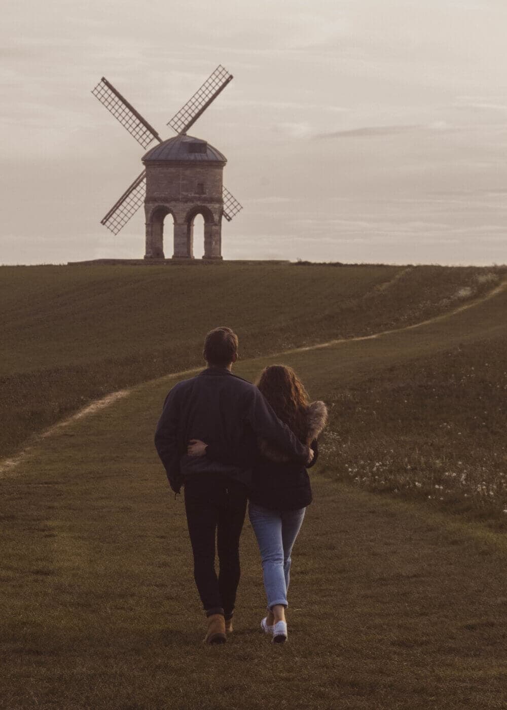 A couple walks arm in arm down a path towards a windmill in the distance under an overcast sky. - Home Instead