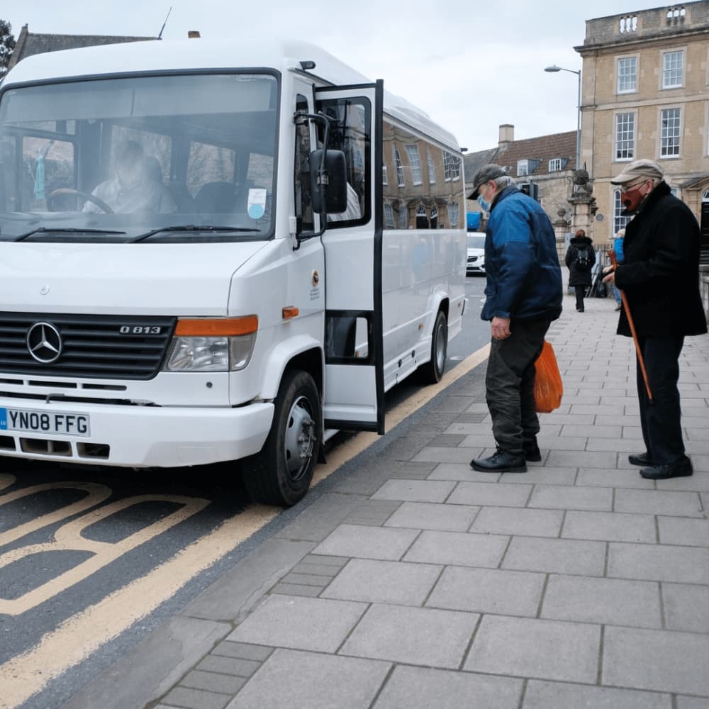 Two elderly men, one with a cane, board a white bus on a city street sidewalk. - Home Instead